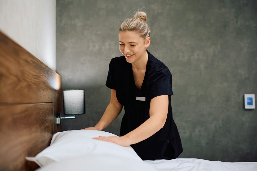 Young happy maid changing bed linen in a hotel room.